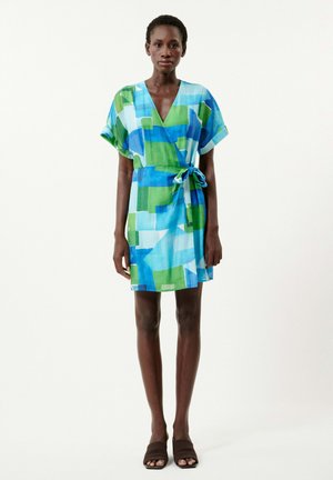 Robe courte en tissu léger avec un motif abstrait bleu et vert, décolleté en V, et une ceinture à la taille. Chaussures plates présentées.