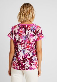 Kvinna med kort blont hår som bär en kortärmad blus med rosa blommor och vita byxor, sedd bakifrån mot en enfärgad bakgrund.