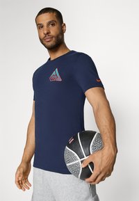 Camiseta de manga corta azul marino con un logo geométrico en el pecho, acento naranja en la manga, acompañada de un balón de baloncesto texturizado en blanco y negro.