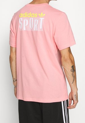 Henkilö pukeutuneena vaaleanpunaiseen Adidas urheilupaitaan, jossa keltavalkoinen logo selässä, mustat housut valkoisilla raitoja ja näkyvät käsivarsitatuoinnit.