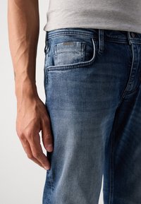 Jeanși din denim albastru închis cu un efect de uzare, design cu cinci buzunare și un eticheta de marcă pe buzunarul din spate. Textura este netedă.