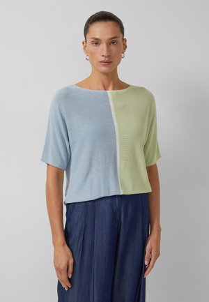 Kobieta w luźnej bluzce z krótkim rękawem z pionowymi, niebieskimi i zielonymi blokami kolorystycznymi, do tego spodnie z wysokim stanem w ciemnym odcieniu niebieskiego.