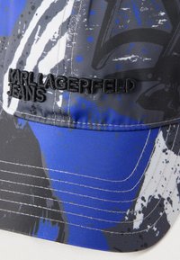 Tuvplāns uz daudzkrāsainu cepuri ar abstraktām zilām, melnām, pelēkām un baltām rakstu zīmēm, uz kuras ir melni izšūts teksts "KARL LAGERFELD JEANS".