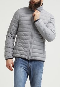 Uomo con barba che sta chiudendo una giacca trapuntata grigio chiaro, indossando jeans blu, in piedi contro uno sfondo semplice e chiaro.