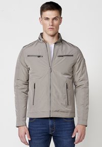 Chaqueta de cremallera gris claro con costuras en los hombros verticales y horizontales, dos bolsillos frontales con cremallera y un cuello alto. Textura suave.