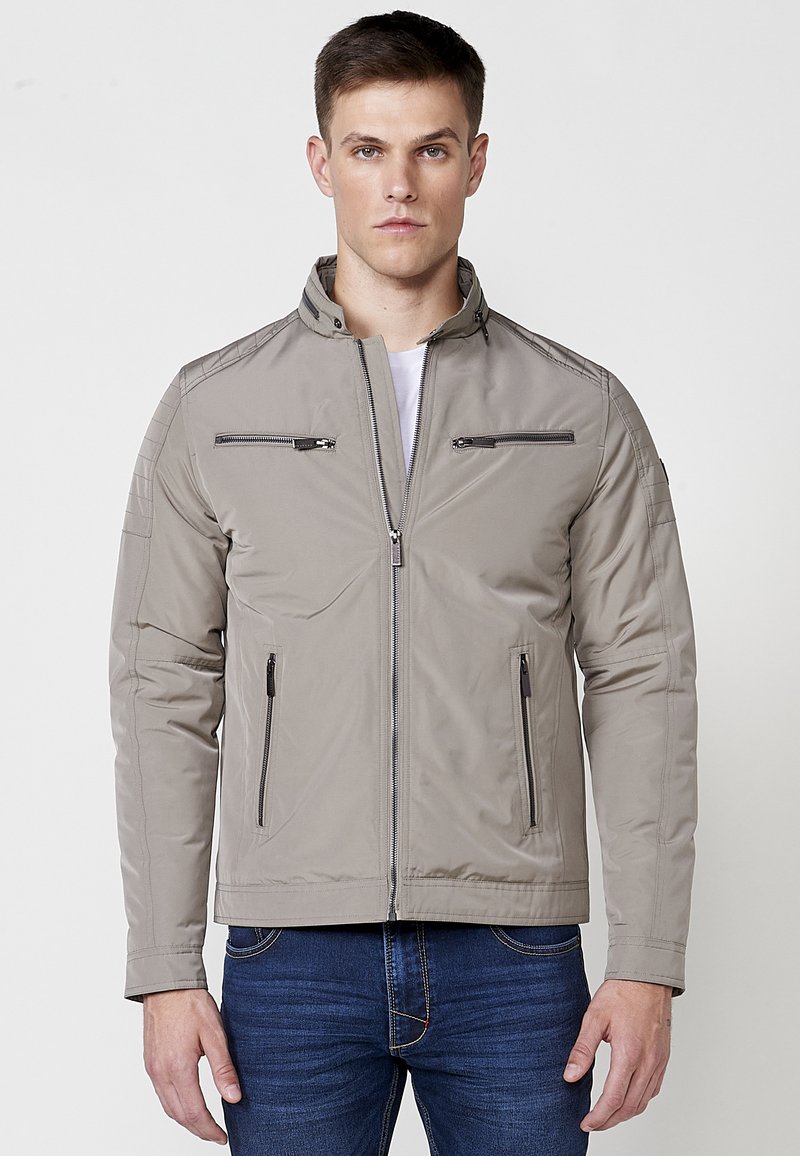 Chaqueta de cremallera gris claro con costuras en los hombros verticales y horizontales, dos bolsillos frontales con cremallera y un cuello alto. Textura suave.