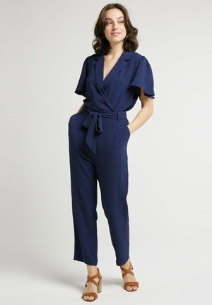 Jumpsuit bleu marine à manches courtes, avec une ceinture à la taille, deux poches latérales et un tissu texturé. Le modèle porte des sandales à talons marron.