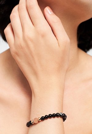 Polso con braccialetto nero di perline con una piccola perla decorativa dorata e una d'argento sulla pelle nuda vicino al collo di una persona.