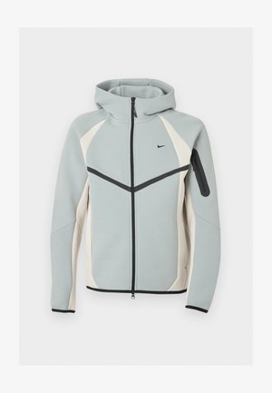 Světlé šedé a bílé zipové bundy s kapucí s černým logem Nike, černým zipem a zipovou kapsou na levém rukávu.