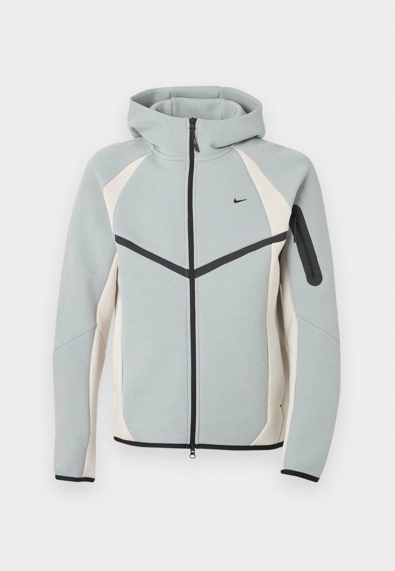 Hellgraue und weiße Kapuzenjacke mit Reißverschluss, schwarzem Nike-Logo, schwarzem Reißverschluss und Reißverschlusstasche am linken Ärmel.