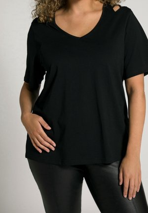 Woman wearing a loose black V-neck T-shirt with a shoulder cutout and black leather pants, hand resting on hip.