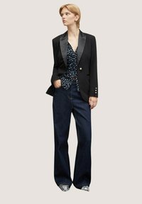 Černé sako se saténovými klopami, modrá vzorovaná blůza a široké tmavé džíny, doplněné stříbrnými botami. Jednoduché pozadí.