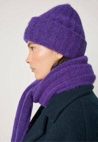 Vijolična volnena kapa in ujemajoči šal, ki imata mehak, teksturiran zaključek. Kapa je rahlo zvita na robu, s prijetnim dizajnom.