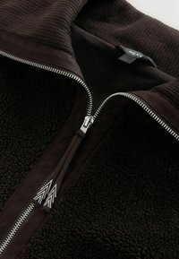 Pruun lukuga jakk, millel on pehme fliisist tekstuur, kontrastne soonikuga krae ja mansetid ning hõbedane tõmblukk dekoratiivse tõmbekeelega.