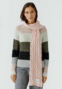 Rosa gestrickter Schal mit einem strukturierten Zopfmuster, getragen �über einem mehrfarbigen gestreiften Pullover. Mit einem Markenetikett an einem Ende.
