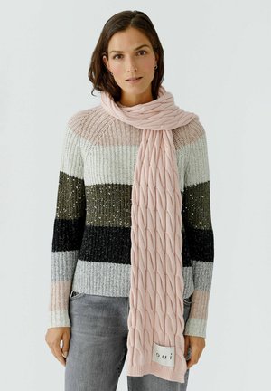 Rosa gestrickter Schal mit einem strukturierten Zopfmuster, getragen über einem mehrfarbigen gestreiften Pullover. Mit einem Markenetikett an einem Ende.