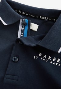 Jūras spēku polos krekls ar struktūru adījumu, baltām un zilu akcentu svītrām, pogu aizdari un logo "BAKER by TED BAKER" priekšā.