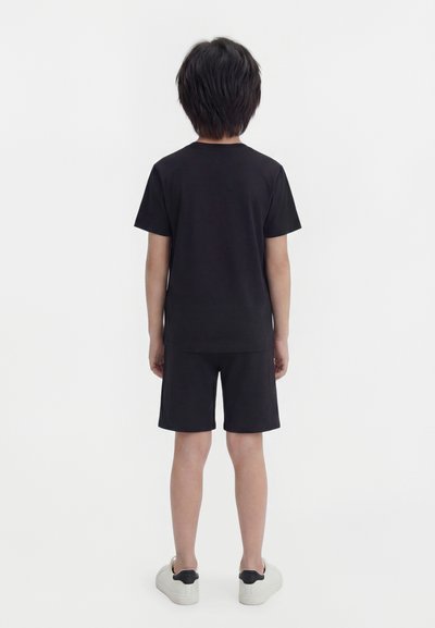 Παιδί που στέκεται με την πλάτη γυρισμένη, φορώντας απλό μαύρο μπλουζάκι, μαύρο σορτς και λευκά αθλητικά παπούτσια με μαύρες λεπτομέρειες στη φτέρνα.