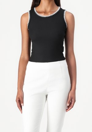 Czarny prążkowany top bez rękawów z okrągłym dekoltem, ozdobiony kontrastującym białym przeszyciem w zygzak na kołnierzu i wokół otworów na ramiona. Połączony z białymi spodniami.