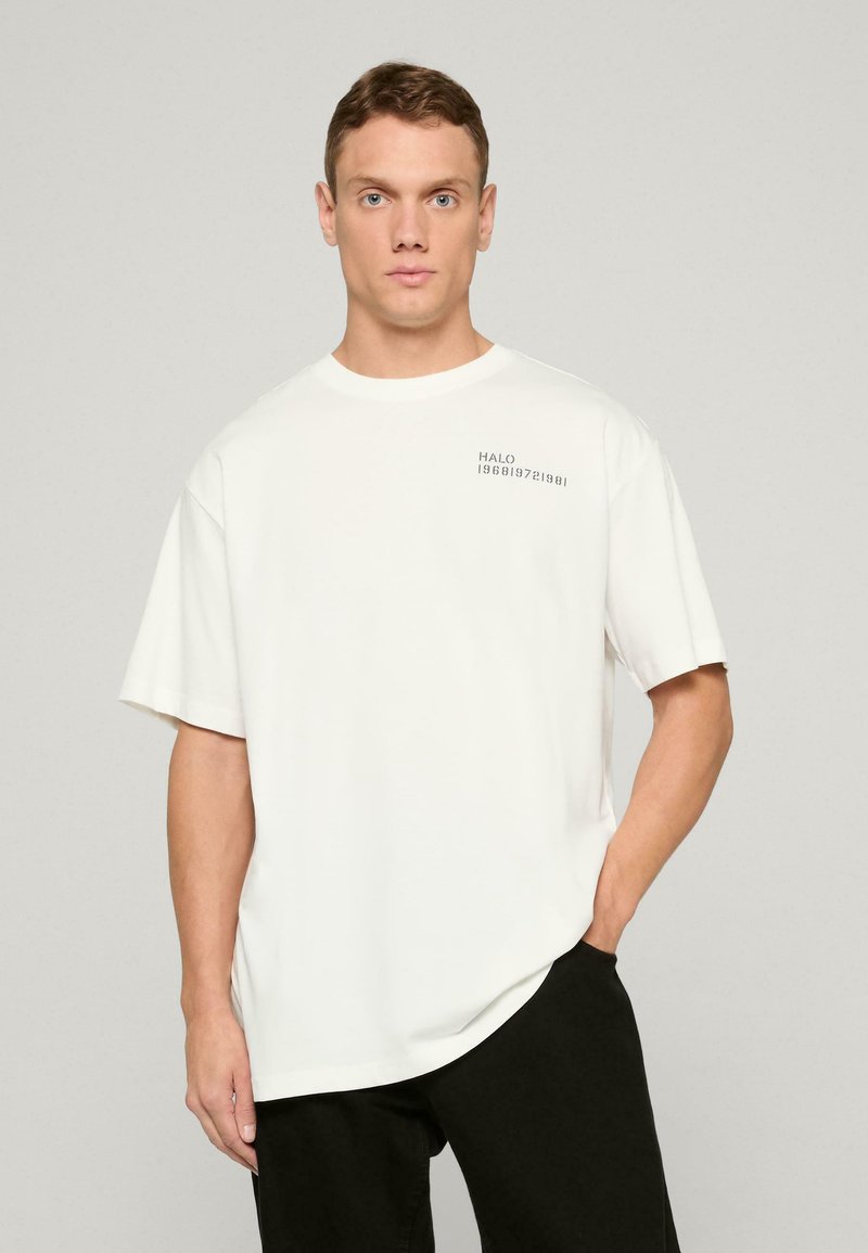 Valkoinen puuvillainen T-paita lyhyillä hihoilla, pyöreällä kaula-aukolla ja hienovaraisella "HALO 196819721981" -printillä vasemmalla puolella. Yhdistetty mustiin housuihin.