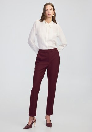 Blouse blanche à manches longues avec détail plissé et col, associée à un pantalon ajusté bordeaux. Le mannequin porte des chaussures à talons hauts bordeaux.