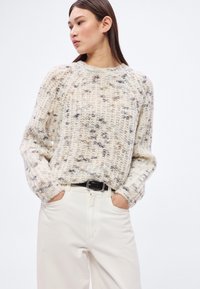 Cremefarbener und grauer Strickpullover mit strukturiertem Muster, weiter Passform und gerippten Bündchen, kombiniert mit weißen, hoch taillierten Hosen und einem schwarzen Gürtel.