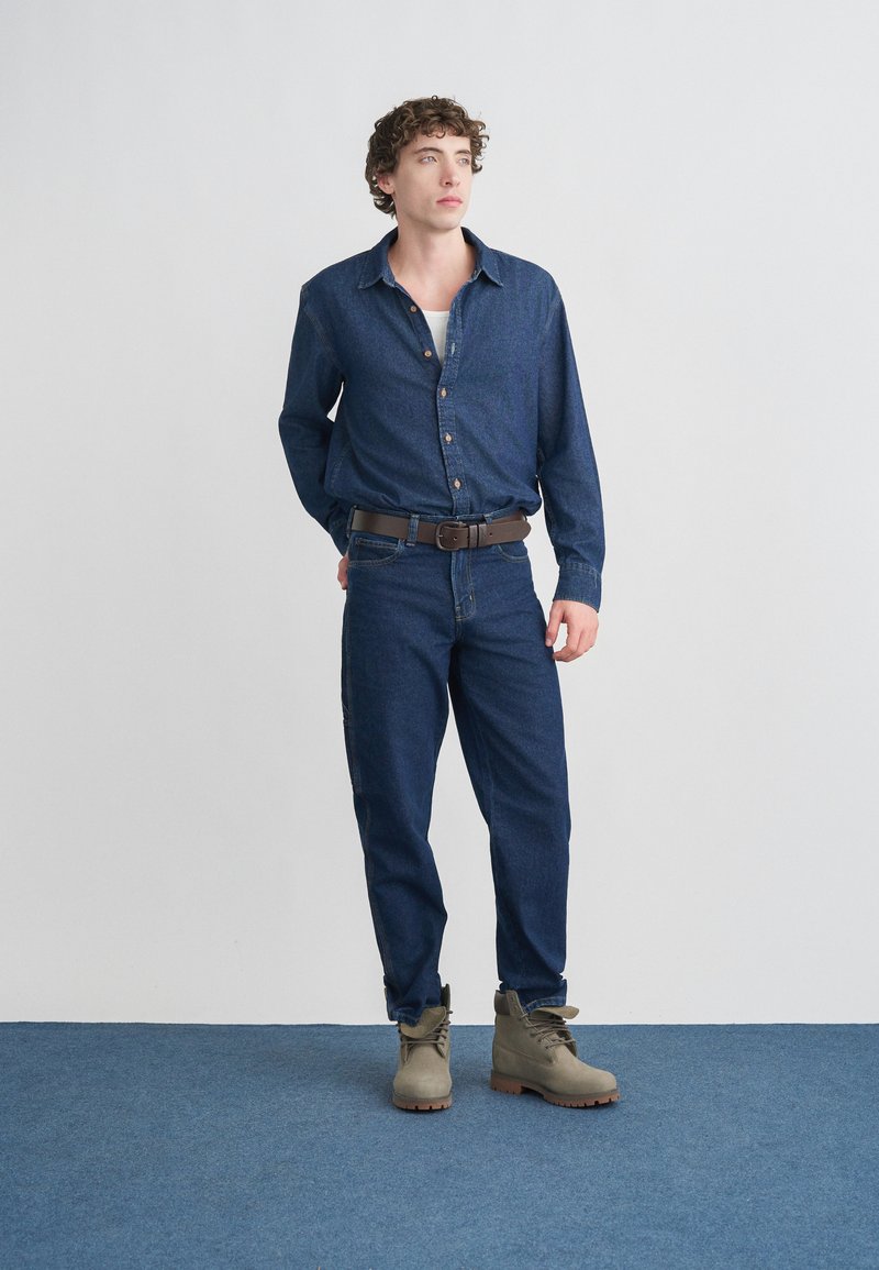 Denim shirt in dark blue, long-sleeved with button front, paired with lighter blue jeans and beige ankle boots with brown soles.