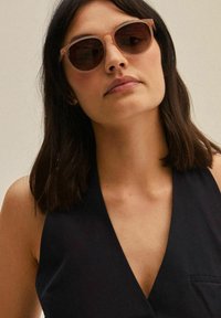 Sunglasses with round, light pink frames and dark lenses. Worn by a person with dark hair, against a neutral background.