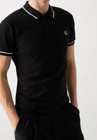 Černé polokošile s bílými akcenty, má límeček, krátké rukávy a logo CK na levé hrudi. Vyrobeno z texturované látky.
