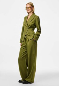Olijfgroene streepjeskostuum met een getailleerde blazer, brede broekspijpen en zwarte schoenen. De look heeft een enkele knoopsluiting en zakken.