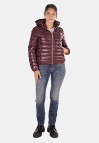 Giacca a vento bordeaux con cappuccio, texture lucida, chiusura con zip e orlo a coste. Abbinata a jeans blu e stivali neri con lacci.