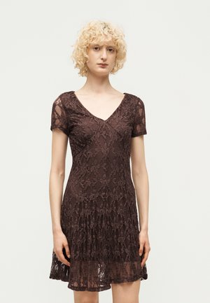 Mujer con cabello rubio rizado que lleva un vestido corto marrón de encaje con cuello en V, de pie frente a un fondo claro y liso.