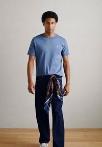 Īsroku zils t-krekls, tumši džinsi un rakstains šalle, kas sasieta ap jostasvietu, veido ikdienišķu, kārtainu izskatu. Gluda auduma tekstūra.