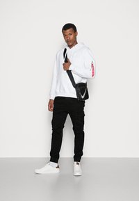 Sudadera blanca con capucha, pantalones cargo negros y zapatillas blancas con detalles en marrón. El modelo posa con un bolso negro de hombro que presenta un logo plateado.
