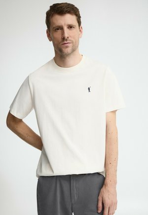 Hombre con cabello corto y castaño, vistiendo una camiseta blanca de manga corta con un pequeño logo negro de jugador de polo y pantalones grises, de pie frente a un fondo blanco.