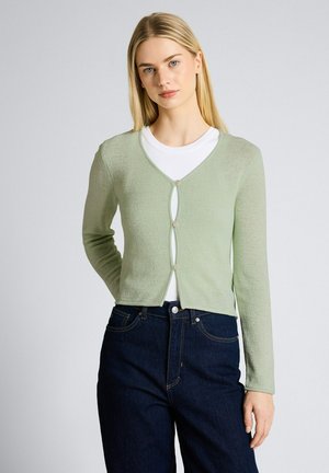 Junge Frau mit langem blondem Haar, die eine hellgrüne, geknöpfte Strickjacke über einem weißen Oberteil und dunkelblauen Jeans trägt und vor einem einfarbigen Hintergrund steht.