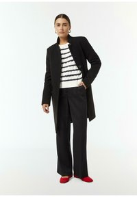 Schwarzer Mantel über einem weißen und marineblauen gestreiften Pullover, kombiniert mit schwarzen Hosen und roten Ballerinas. Glattes Material und maßgeschneiderter Design.