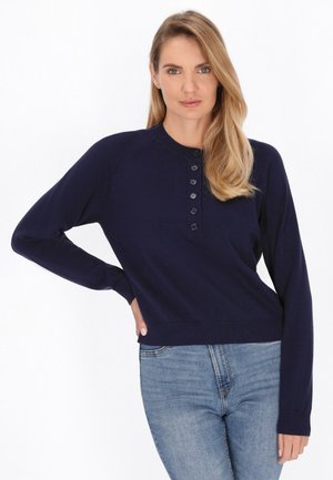Frau mit blondem Haar, die einen marineblauen, geknöpften Pullover und blaue Jeans trägt, steht mit einer Hand auf der Hüfte vor weißem Hintergrund.