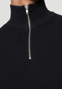 Pull noir en maille avec un col montant muni d'une fermeture éclair argentée. La texture du tissu est lisse et tissée serrée.