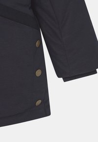 Tmavě námořnická bunda s rukávem, na kterém jsou tři bronzové patentky podél strany a žebrovaný manžeta na zápěstí.