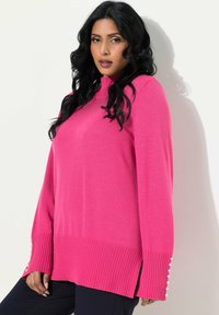 Pull en rose avec un col montant, une finition côtelée en bas et des fentes sur les côtés. Présente des boutons décoratifs en perles sur les manches. Tissu tricoté doux.