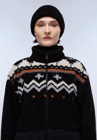 Giacca in pile nera con zip e design a motivi in bianco, grigio e arancione. Presenta un alto colletto e un materiale morbido e strutturato. Indossata con un berretto.