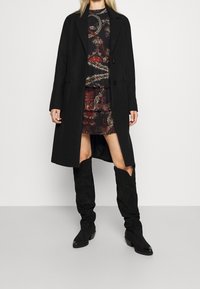 Manteau en laine noire à coupe droite, avec deux boutons sur le devant et des poches latérales, porté sur une robe à motifs et des bottes en daim noir montant jusqu'aux genoux.