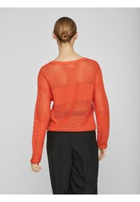 Röd stickad tröja med öppet vävd design, med en bred halsringning och långa ärmar. Bärs över svarta byxor, vilket visar texturerade detaljer.