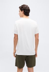 Άνδρας με την πλάτη, φοράει απλό λευκό κοντομάνικο πουκάμισ ο και σκούρα πράσινα σορτς, σε ανοιχτό φόντο.