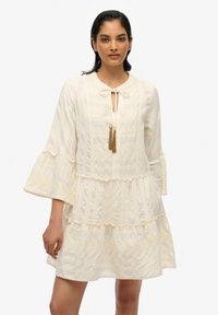 Cremfarbenes, schichtweise gestaltetes Kleid mit einem runden Ausschnitt und einerBindedetail, ausladenden Ärmeln und einem strukturierten Muster. Akzentuiert mit goldenen Quasten.