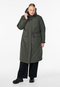 Ženska prilagaja kapuco dolgemu, olivnozelenemu parkastemu plašču, oblečena v črne široke hlače in črne škornje, stoji na beli podlagi.