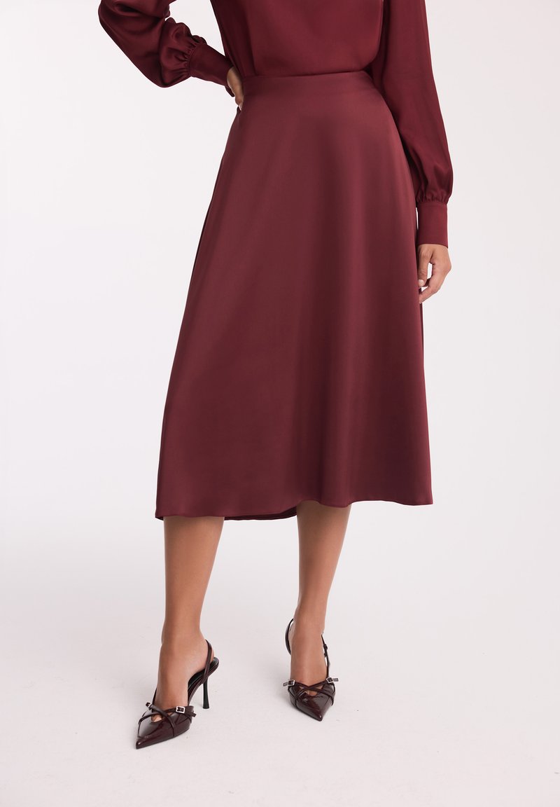 Burgundowa satynowa spódnica midi o gładkiej fakturze i rozszerzanym kroju, w połączeniu z ciemnymi, zwężanymi szpilkami typu slingback z akcentem w postaci kokardy.