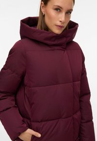 Vínová prošívaná bunda s vysokým límcem, prošívaným designem a bočními kapsami. Hladká textura s moderním, strukturovaným tvarem.