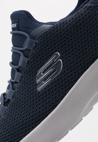 Marineblaues Mesh-Sneaker mit strukturiertem Obermaterial, ausgestattete mit reflektierendem Logo, elastischen Schnürsenkeln und einer weißen Gummisohle. Leicht und atmungsaktiv im Design.